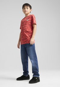 Raudona grafika marškiniai su juodais spaudiniais, mėlyni džinsai ir juodi sportbačiai. Tiesaus silueto džinsai, trumpomis rankovėmis ir logotipo akcentais ant marškinėlių.