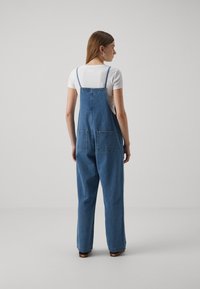 Denim kombinezon svetlo modre barve z nastavljivimi naramnicami, dvema velikima zadnjima žepoma in sproščenim krojem, v kombinaciji z oprijeto belo majico.