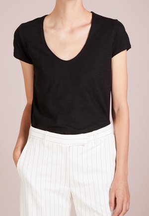 Vrouw draagt een zwart T-shirt met korte mouwen en een V-hals, ingestopt in witte hoge taillebroek met dunne verticale krijtstrepen, handen in de zakken.