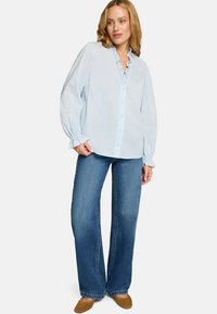 Blouse bleu clair à manches longues avec col à volants et fermeture à boutons, associée à un jean denim à jambes larges et des chaussures marron sans lacets.