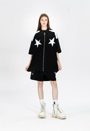 Joven mujer con camisa negra de cremallera grande con estrellas blancas, pantalones cortos negros, calcetines blancos y botines blancos con cordones, de pie sobre fondo blanco.