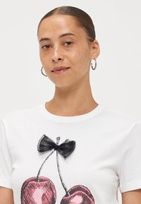 T-shirt en coton blanc avec une illustration de cerises roses, agrémentée d'un nœud noir transparent au-dessus des cerises et d'un col rond.