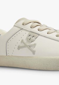 Baskets en cuir blanc avec un détail de crâne et os croisés gris, bout rond et semelle en caoutchouc texturée. Lacets beige et accents perforés sur les côtés.