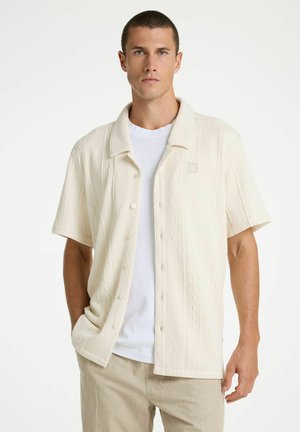 Jonge man met kort haar, gekleed in een gestructureerd, off-white kortermig overhemd over een wit T-shirt en beige broek, rechtopstaand.