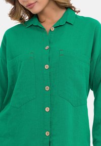 Groene linnen shirt met een kraaghalslijn, voorzien van houten knopen, twee voorzakken en stikaccenten.