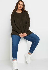 Kakifärgad stickad tröja med ribbad textur, tillsammans med blå skinny jeans och vita sneakers. Modell sittande på en vit block.