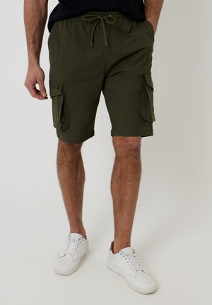 Uomo che indossa pantaloncini cargo verde oliva con coulisse e sneakers bianche, in piedi con le mani nelle tasche su sfondo semplice.