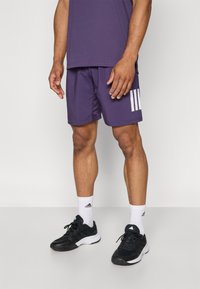 Lila idrottsshorts med vitt tre-stripes logotyp, kombinerat med svarta sneakers och vita ankelsockor. Tyget är lätt och slätt.