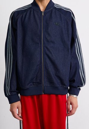 Persona que lleva una chaqueta azul oscuro con cremallera y rayas azul claro en las mangas y pantalones rojos, visible desde el cuello hasta las caderas.