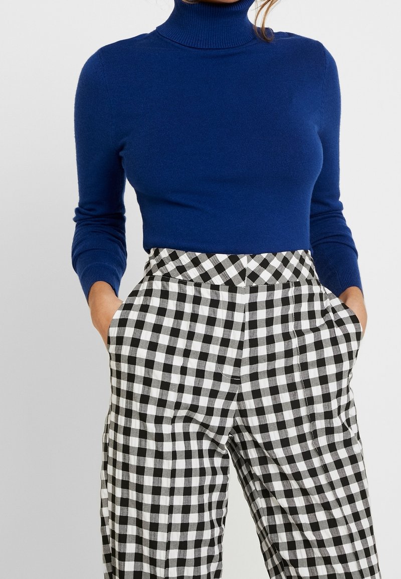 Maglione a collo alto blu realizzato in morbido tessuto a maglia, abbinato a pantaloni a quadri bianchi e neri a vita alta, dotati di tasche laterali.