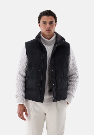 Mann trägt schwarze gesteppte Weste über hellgrauem strukturiertem Rollkragenpullover und weißen Hosen, steht vor einfachem Hintergrund.