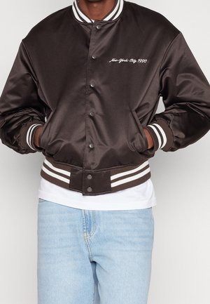 Personne portant une veste bomber en satin marron foncé avec des poignets et un col côtelés rayés blancs, et un jean bleu clair.