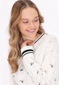 Pulover alb tricotat cu guler și manșete negre din tricot, având broderie florală colorată și un model texturat.