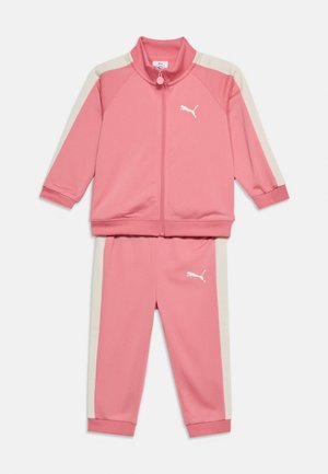 Survêtement pour enfant en deux pièces rose avec bandes blanches sur les côtés, veste zippée intégrale, poignets élastiques, et logo Puma sur la veste et le pantalon.