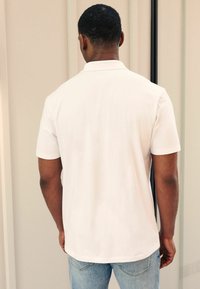 Camiseta polo blanca de manga corta, con textura suave, diseño con cuello y dobladillo recto, usada con jeans de color azul claro. Vista desde la parte posterior.