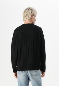 Sort ribbestrikket sweater med en afslappet pasform, rund halsudskæring og lange ærmer. Strikket tekstur, minimal branding ved sømmen, båret med lyseblå jeans.
