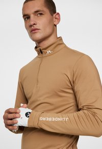 Długosięgnący, zapinany na zamek top w tanowym materiale o gładkiej fakturze. Posiada wysoki kołnierz i biały akcent z logo na rękawie.