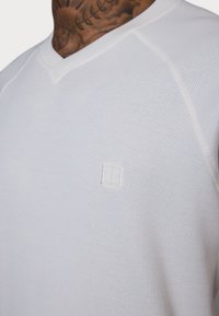 Weißes, strukturiertes Langarmhemd mit V-Ausschnitt, versehen mit einem kleinen gestickten Logo auf der Brust und kontrastierenden Nähten entlang der Ärmel.