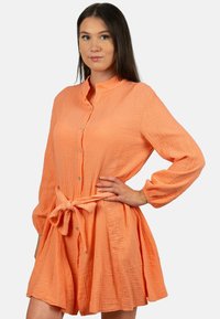 Abito camicia color pesca realizzato in tessuto testurizzato, con maniche lunghe, colletto alla mandarino, bottoni frontali e un dettaglio in vita legato.
