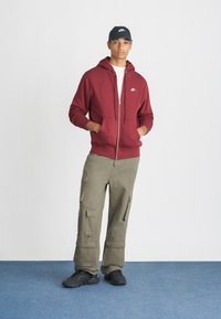 Sweatshirt marrom com fecho, com bolsos tipo canguru, combinado com calças cargo verdes-oliva com múltiplos bolsos laterais, e sapatilhas pretas.