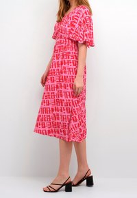 Pink dress featuring a geometric pattern in varying shades of red, with a V-neck, short sleeves, and a flowy, knee-length design.