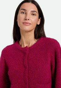 Fuchsia strukturierte Strickjacke mit Rundhalsausschnitt, vorderem Knopfverschluss und zwei Taschen an der Taille. Aus weichem Wollmix-Material.