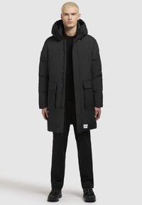 Črn puhasti plašč s kapuco, z gumbi in zadrgo, dvema sprednjima žepoma ter oznako na robu. Gladek material in dolga silhueta.