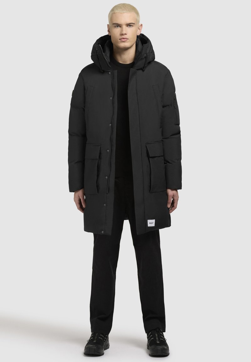 Črn puhasti plašč s kapuco, z gumbi in zadrgo, dvema sprednjima žepoma ter oznako na robu. Gladek material in dolga silhueta.