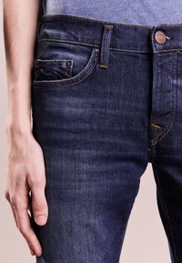 Mörkblå denimjeans med rak skärning, framträdande sömmar och en liten myntficka fram. Synlig metallknappsstängning.