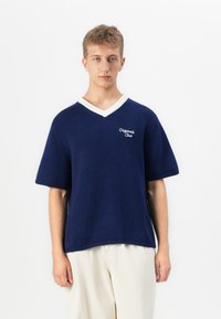 Jeune personne avec des cheveux châtain clair courts portant un pull bleu marine à col en V et à manches courtes avec des bords blancs, associé à un pantalon large crème, le tout devant un fond blanc.