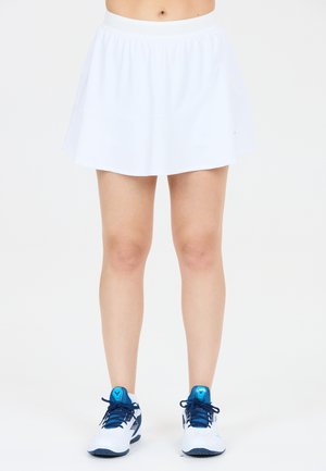 Kvinde iført hvid tennisnederdel og hvide atletik sko med blå snørebånd, stående mod en ensfarvet baggrund.