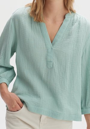 Vrouw draagt een losse lichtgroene blouse met een gestructureerd patroon en driekwartmouwen en een beige broek, hand in zak.