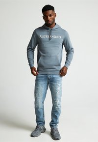 Sudadera con capucha gris con "ALESSANDRO" impreso en blanco, combinada con jeans claros desgastados y sneakers grises.