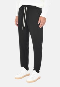 Persona che indossa pantaloni neri con coulisse, una maglietta bianca a maniche lunghe, calze nere e sneakers bianche con suole color gomma, in piedi contro uno sfondo bianco.