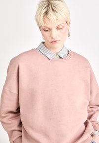 Felpa rosa chiaro con scollatura rotonda e spalle cadute. Sotto, è visibile una camicia a righe blu e bianche con bottoni.