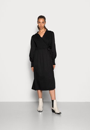 Vestido preto envolvente com mangas longas e corpete texturizado, combinado com botins bicolores com bico fino e salto baixo.
