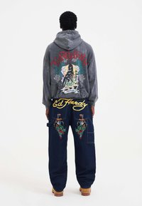 Grå hættejakke med farverig grafik på bagsiden, kombineret med blå denimbukser med livlige broderede designs og en afslappet pasform.