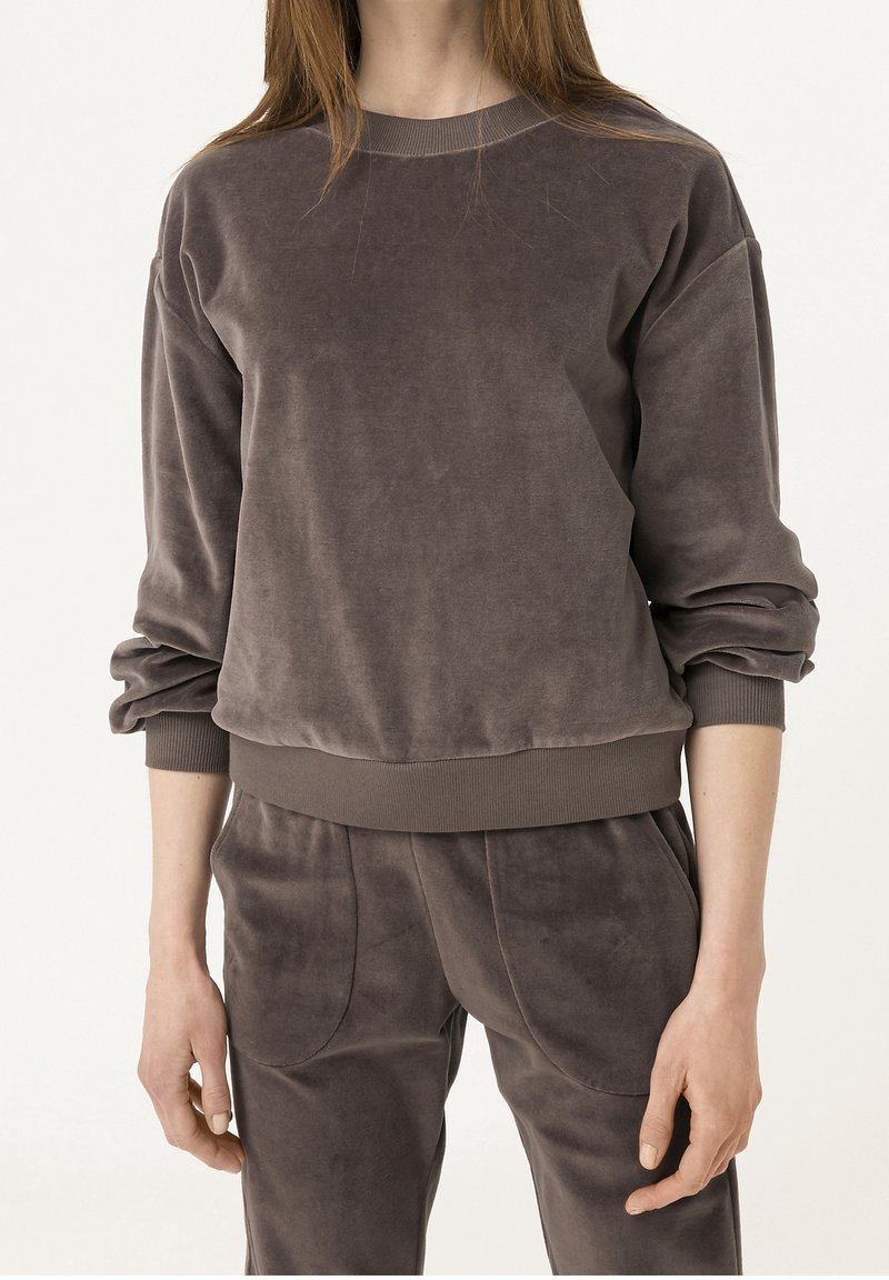 Kobieta ubrana w brudnoróżową welurową bluzę z długim rękawem oraz dopasowane welurowe spodnie z wysokim stanem i elastycznym paskiem.