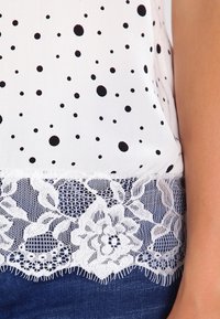 Blusa blanca con lunares negros, con un borde de encaje en el canto inferior. El encaje tiene patrones florales y es festoneado.