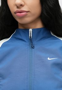 Veste Nike en tissu bleu avec une fermeture éclair et un logo blanc. Présente un col montant et des accents contrastants le long des épaules. Texture lisse.