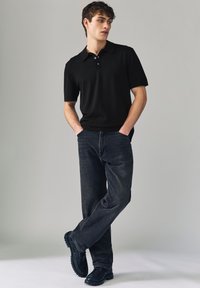 Polo noir à manches courtes avec col classique, associé à un jean en denim foncé et des chaussures noires à semelles texturées. Apparence simple et moderne.