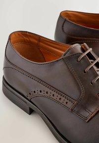 Zapatos de vestir de cuero marrón con un acabado suave, que presentan detalles perforados y cordones. El interior es de cuero beige. Suela de goma para mayor adherencia.