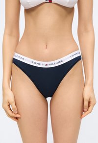 Γυναίκα που φοράει εσώρουχα Tommy Hilfiger σε ναυτικό μπλε με λευκή μέση και λογότυπο, εμφανιζόμενη από τη μέση μέχρι το μέσο του μηρού με φόντο απλό.