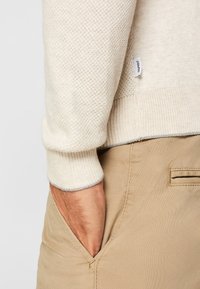 Pull en tricot beige clair avec manches texturées, doté d'un poignet côtelé. Main dans la poche d'un pantalon chino en coton tan, affichant une étiquette de marque discrète.