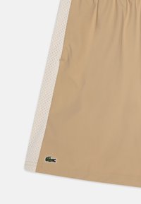 Shorts beiges avec des panneaux latéraux en mesh, taille élastique et logo de crocodile vert brodé près de l'ourlet. Texture lisse, design décontracté.