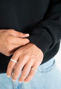 Persona che sistema il polsino di un maglione nero, con anelli d'argento sulle dita, mentre nel fondo si vedono dei jeans azzurri chiari.
