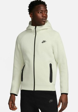 Felpa con zip Nike in tessuto verde chiaro dalla texture liscia. Presenta un logo nero, tasche con zip e un cappuccio aderente.