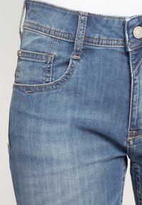 Närbild av blå denimjeans som visar framfickan, bältesöglan och knappen med synliga söm detaljer.