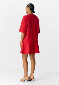 Abito rosso strutturato con maniche corte e gonna svasata. Il materiale sembra liscio, con un dettaglio di cucitura sulla schiena. Indossato con tacchi di colore chiaro.