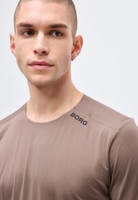 Νεαρός άνδρας με κοντά μαλλιά, φορώντας μια γκριζοκαφέ μπλούζα με στρογγυλή λαιμόκοψη και την επιγραφή "BORG" κοντά στον ώμο, στραμμένος ελαφρώς προς τα αριστερά μπροστά σε απλό φόντο.